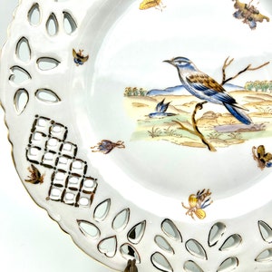 May include: White porcelain decorative plate with a pierced edge and a painted bird and butterfly design. The plate features a detailed illustration of a blue and brown bird perched on a branch, with several butterflies scattered around the rim.