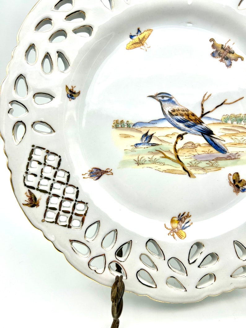 May include: White porcelain decorative plate with a pierced edge and a painted bird and butterfly design. The plate features a detailed illustration of a blue and brown bird perched on a branch, with several butterflies scattered around the rim.