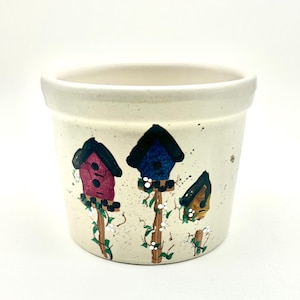May include: Cream-colored ceramic flower pot with hand-painted birdhouse design. The birdhouses are in shades of red, blue, and yellow, with green vines and white accents. The pot has a wide opening and a slightly textured surface.