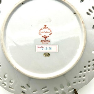 May include: A white porcelain plate with a decorative, pierced edge. The underside displays a red and gold emblem, a sticker with "Item No. 14-45416", and a "Made in China" sticker. The plate is held by a bronze-coloured stand.