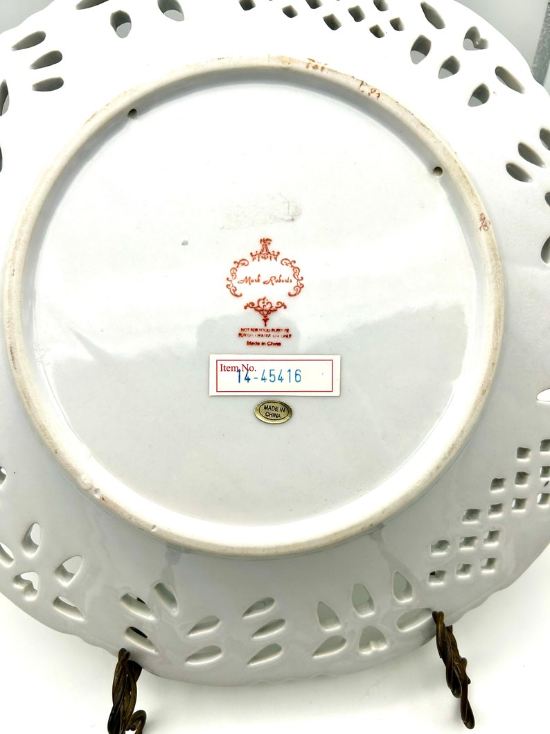 May include: A white porcelain plate with a decorative, pierced edge. The underside displays a red and gold emblem, a sticker with "Item No. 14-45416", and a "Made in China" sticker. The plate is held by a bronze-coloured stand.