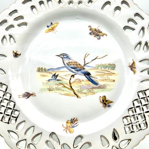 May include: White porcelain decorative plate with a pierced rim and a painted bird and butterfly design. The plate features a central image of a blue and brown bird perched on a branch, surrounded by colourful butterflies.