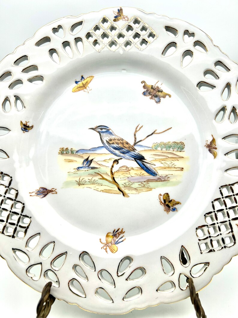 May include: White porcelain decorative plate with a pierced rim and a painted bird and butterfly design. The plate features a central image of a blue and brown bird perched on a branch, surrounded by colourful butterflies.