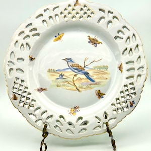 May include: White porcelain decorative plate with a pierced rim and gold trim. The plate features a painted scene of a blue and brown bird perched on a branch, with butterflies and a landscape background. The plate is displayed on a brass stand.