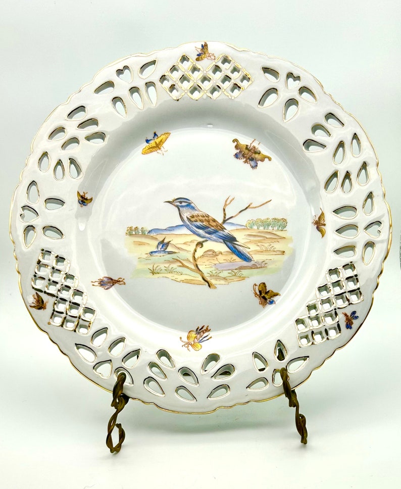 May include: White porcelain decorative plate with a pierced rim and gold trim. The plate features a painted scene of a blue and brown bird perched on a branch, with butterflies and a landscape background. The plate is displayed on a brass stand.