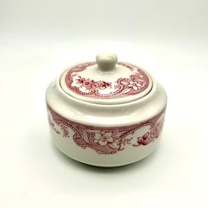 以下が含まれることがあります： 赤い花柄の白い陶器製の砂糖入れ。蓋はノブ付きです。