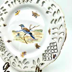 May include: White porcelain decorative plate with a pierced rim and gold trim. The plate features a central image of a blue and brown bird perched on a branch, with butterflies and a landscape scene. The plate is displayed on a decorative metal stand.