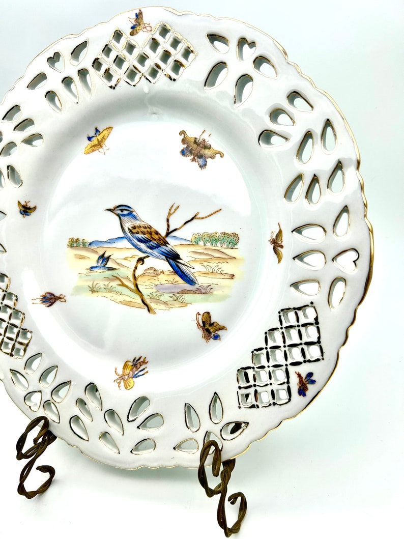 May include: White porcelain decorative plate with a pierced rim and gold trim. The plate features a central image of a blue and brown bird perched on a branch, with butterflies and a landscape scene. The plate is displayed on a decorative metal stand.