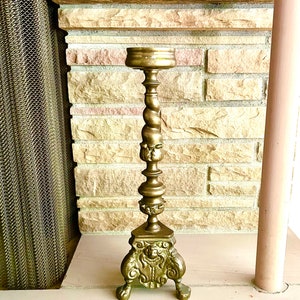 May include: A gold metal candlestick with a twisted stem and a decorative base with three feet. The candlestick is in front of a brick wall.