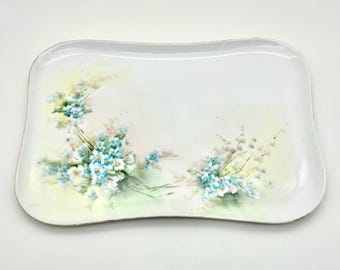 May include: A rectangular white ceramic tray with curved edges, featuring a hand-painted design of clusters of small light blue flowers with green stems and leaves, accented by soft yellow and pale green watercolor-style washes.