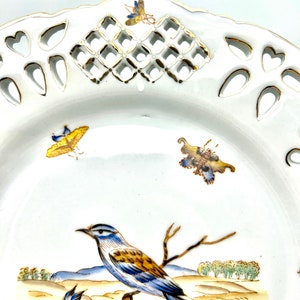 May include: White porcelain decorative plate with a detailed bird and landscape scene. The plate features a pierced rim with heart and diamond-shaped cutouts, and is accented with gold trim and colourful insect illustrations.
