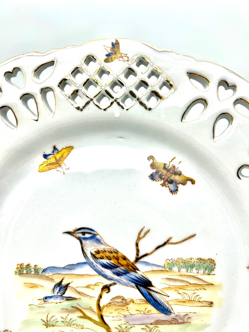 May include: White porcelain decorative plate with a detailed bird and landscape scene. The plate features a pierced rim with heart and diamond-shaped cutouts, and is accented with gold trim and colourful insect illustrations.