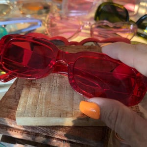 May include: Rectangular sunglasses with a translucent red plastic frame and matching red tinted lenses. The eyewear features a simple, streamlined design with a smooth finish, showcasing a vibrant, solid red color throughout the entire frame and lens structure.