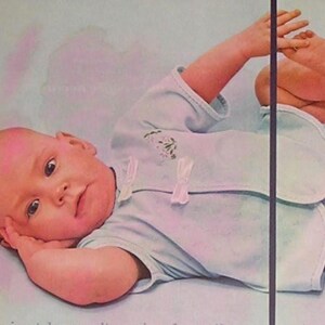 Puede incluir: Un beb&eacute; con un body azul claro con ribete blanco y un babero, acostado sobre una superficie azul claro. El beb&eacute; mira al espectador con una mano cerca de su cara y el otro brazo y pierna levantados.