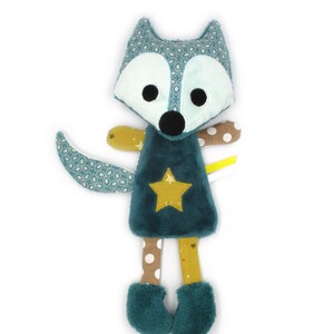 Peut inclure: Une poupée renard en peluche turquoise avec une tête et une queue à motifs, un visage blanc et des yeux noirs. Elle porte une robe turquoise avec une étoile dorée, des jambes marron et jaunes et des bottes en fourrure turquoise. Le texte "Mélomélie création" est en haut.