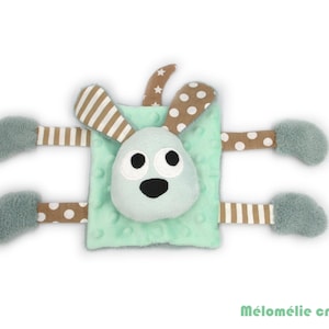 May include: A mint green baby comforter with a bunny face and four appendages. The bunny face has large black eyes and a black nose. The ears and appendages have brown and white striped and polka dot patterns. The text "Mélomélie création" is in the bottom right corner.