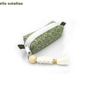 Peut inclure: Une petite pochette rectangulaire à imprimé floral vert, avec une fermeture éclair blanche et un porte-clés à pompon blanc. La pochette a une languette vert foncé à une extrémité. Le texte "Mélomélie création" est en haut.
