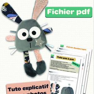 Puede incluir: Un juguete de peluche de conejo gris y rosa con una estrella en el pecho. El juguete viene acompañado de un patrón PDF e instrucciones para hacer el juguete. El texto "Patron doudou lapin" y "Fichier pdf" es visible en la imagen. El texto "Tuto explicatif avec photos" también es visible en la imagen.