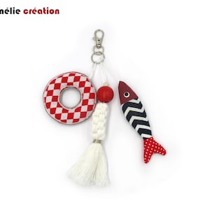 Peut inclure: Un porte-clés composé d'une bouée de sauvetage à carreaux rouges et blancs, d'une perle en bois rouge, d'un cordon en macramé blanc avec un pompon, et d'un charm en forme de poisson à chevrons noirs et blancs avec une queue à pois rouges.