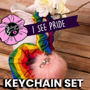May include: A rainbow-colored scrunchie keychain set with a pink fluffy pom-pom charm. The scrunchie has the text "I SEE PRIDE" on a black ribbon. The keychain is labeled "KEYCHAIN SET".