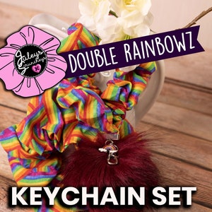 May include: A set of two rainbow-striped scrunchies and a burgundy faux fur keychain with a rainbow charm. The scrunchies and keychain are displayed on a wooden surface. The text "DOUBLE RAINBOWZ KEYCHAIN SET" is visible in the image.