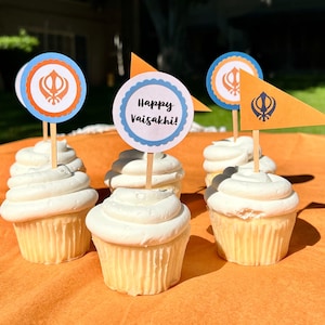 Puede incluir: Seis cupcakes blancos con decoraciones azules y naranjas en la parte superior. Las decoraciones dicen "Happy Vaisakhi!" y presentan un símbolo. Los cupcakes están sobre un mantel naranja.