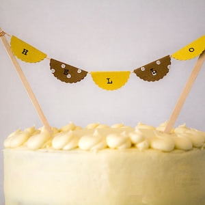 May include: A white frosted cake with a yellow and brown paper banner that reads "HELLO" on top.