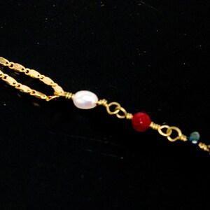 May include: A gold chain necklace with a white pearl, red beads, and a blue bead. The necklace is on a black background.