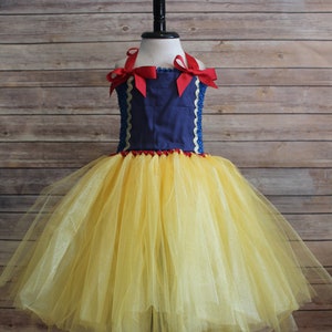 May include: A Snow White-inspired costume dress. The bodice is navy blue with red trim and white lace accents. The skirt is a full, fluffy yellow tulle. Red bows adorn the shoulders. The dress is displayed on a black mannequin.