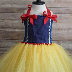 May include: A Snow White-inspired tutu dress. The bodice is navy blue with red ribbon straps and bows. The skirt is yellow tulle. Gold trim accents the bodice. The dress is displayed on a mannequin.