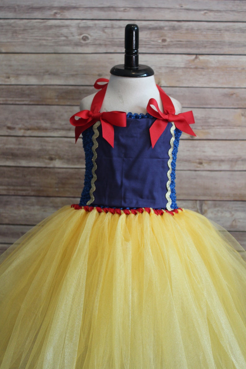 May include: A Snow White-inspired tutu dress. The bodice is navy blue with red ribbon straps and bows. The skirt is yellow tulle. Gold trim accents the bodice. The dress is displayed on a mannequin.