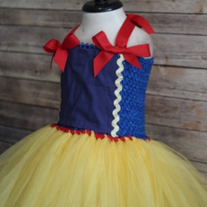 May include: A Snow White-inspired tutu dress with a blue bodice and a yellow tulle skirt. The bodice features red ribbon straps and a white scalloped trim. The dress is displayed on a mannequin.
