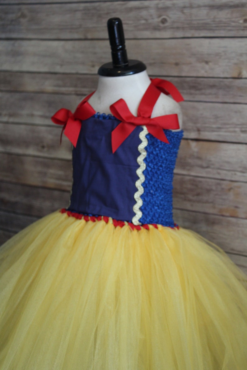 May include: A Snow White-inspired tutu dress with a blue bodice and a yellow tulle skirt. The bodice features red ribbon straps and a white scalloped trim. The dress is displayed on a mannequin.