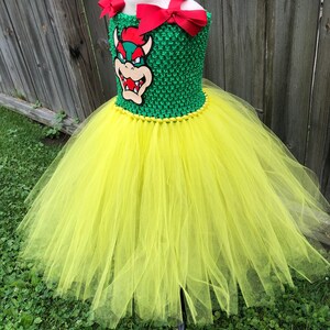 Puede incluir: Un vestido de tut&uacute; de tul amarillo con un corpi&ntilde;o verde que presenta un personaje de dibujos animados con una concha roja y un lazo rojo.