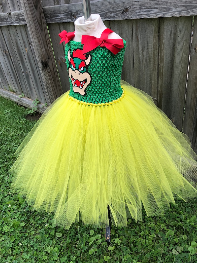 Puede incluir: Un vestido de tut&uacute; de tul amarillo con un corpi&ntilde;o verde que presenta un personaje de dibujos animados con una concha roja y un lazo rojo.