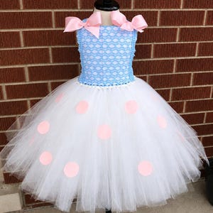 May include: A dress featuring a light blue bodice with a white lace overlay and two large light pink bows at the shoulders. The skirt is made of layers of white tulle decorated with light pink circular polka dots and a white pearl-like trim.