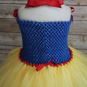 May include: A Snow White-inspired costume featuring a blue crocheted bodice with red ribbon trim and bows. The skirt is made of yellow tulle. A large red bow adorns the neckline.