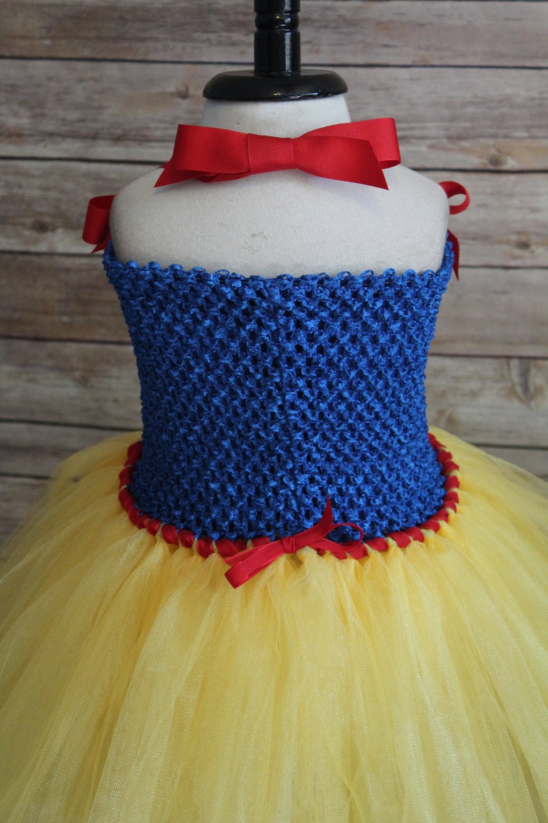 May include: A Snow White-inspired costume featuring a blue crocheted bodice with red ribbon trim and bows. The skirt is made of yellow tulle. A large red bow adorns the neckline.