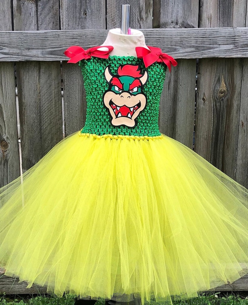 Puede incluir: Una falda de tut&uacute; de tul amarillo con un corpi&ntilde;o verde que presenta una imagen de dibujos animados de Bowser de la serie de videojuegos Super Mario Bros. El corpi&ntilde;o tiene lazos rojos en las correas.