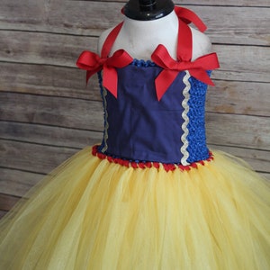 May include: A Snow White-inspired tutu dress with a navy blue bodice, red ribbon straps, and a yellow tulle skirt. The bodice is trimmed with gold and red accents, and the dress is displayed on a mannequin.
