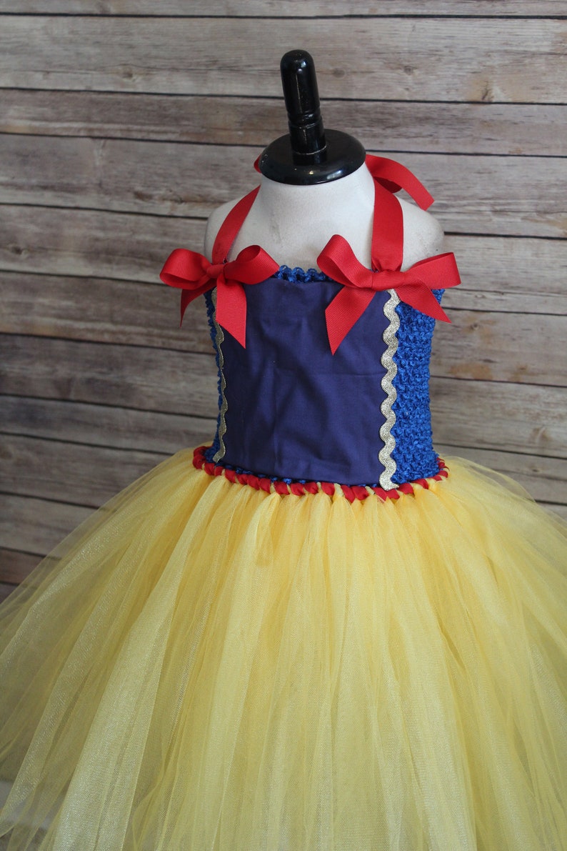 May include: A Snow White-inspired tutu dress with a navy blue bodice, red ribbon straps, and a yellow tulle skirt. The bodice is trimmed with gold and red accents, and the dress is displayed on a mannequin.