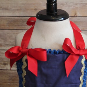 May include: A navy blue dress with red ribbon shoulder ties and bows. The dress has a blue crocheted neckline with gold trim. The dress is displayed on a mannequin.