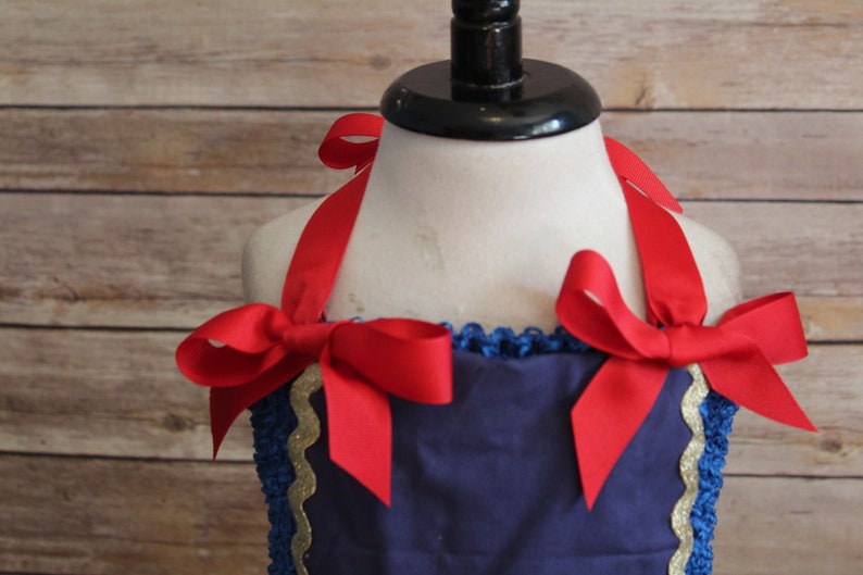 May include: A navy blue dress with red ribbon shoulder ties and bows. The dress has a blue crocheted neckline with gold trim. The dress is displayed on a mannequin.