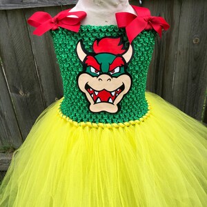 Puede incluir: Un vestido de tut&uacute; verde y amarillo con un lazo rojo en cada hombro. El corpi&ntilde;o presenta una tela verde de crochet con una imagen de dibujos animados de Bowser del videojuego Super Mario Bros.
