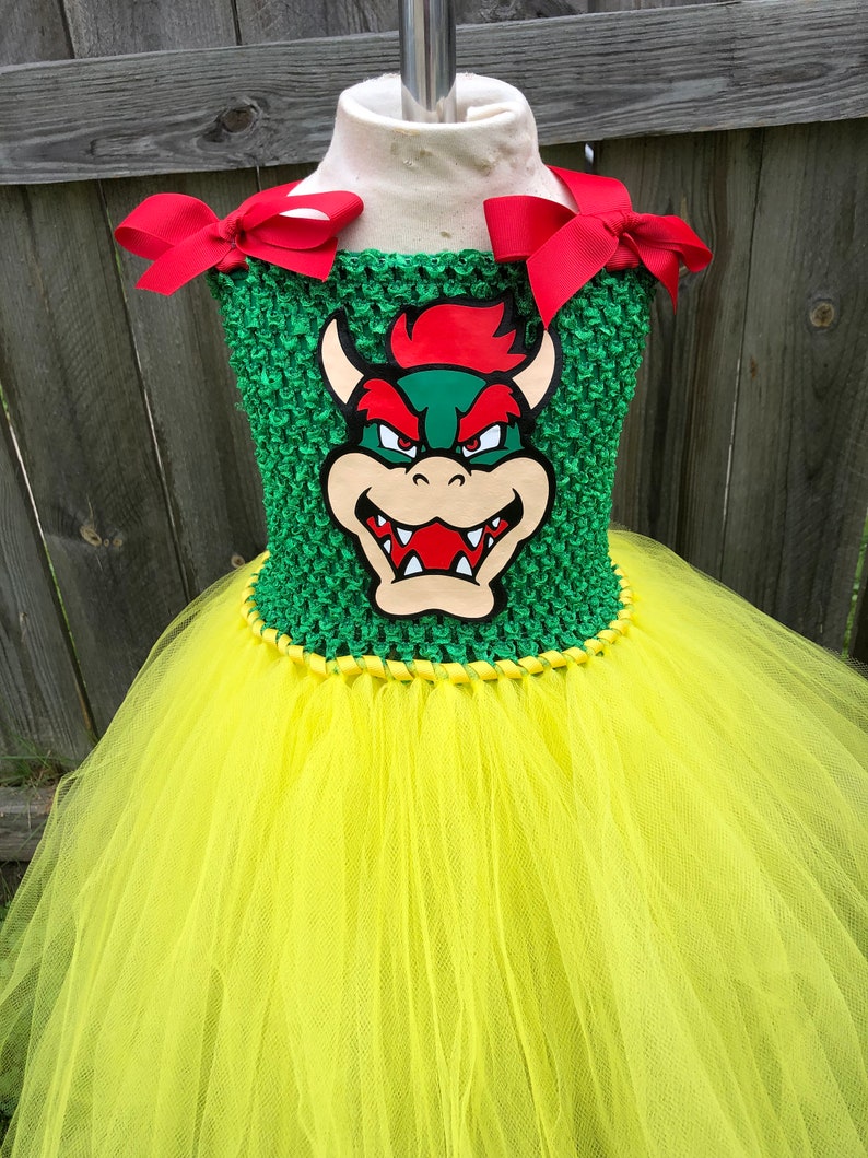 Puede incluir: Un vestido de tut&uacute; verde y amarillo con un lazo rojo en cada hombro. El corpi&ntilde;o presenta una tela verde de crochet con una imagen de dibujos animados de Bowser del videojuego Super Mario Bros.