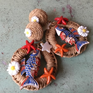 Peut inclure: Une paire de boucles d'oreilles en paille tressée avec des breloques de poissons bleus et rouges, des marguerites blanches, des étoiles de mer orange et rouges et des breloques d'étoiles blanches.