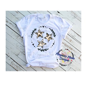 Puede incluir: Camiseta blanca con un diseño en blanco y negro de tres estrellas en un círculo. Las estrellas están rellenas con un estampado de leopardo. El diseño está rodeado de una corona de hojas y corazones.
