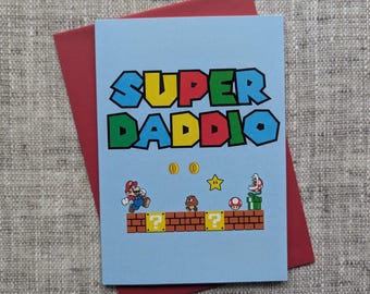 Puede incluir: Una tarjeta de felicitación azul claro con el texto SUPER DADDIO en una fuente pixelada multicolor. La tarjeta presenta una escena de videojuego con Mario, un Goomba, un hongo, una estrella, dos monedas de oro y una planta Piraña sobre ladrillos.