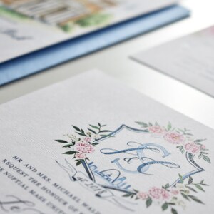 May include: A wedding invitation with a blue and white watercolor design. The invitation features a crest with the initials "J & E" surrounded by pink flowers and green leaves. The text "Mr. and Mrs. Michael Walsh Request the honour of your presence at the nuptial mass united in holy matrimony Saturday, the 27th of October, 2018 at 4:00 p.m.  St. Augustine's Church,  followed by a reception at The  Carolina Inn Chapel Hill, North Carolina" is printed on the invitation.