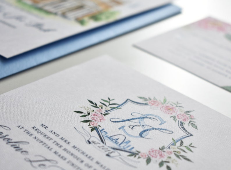 May include: A wedding invitation with a blue and white watercolor design. The invitation features a crest with the initials "J & E" surrounded by pink flowers and green leaves. The text "Mr. and Mrs. Michael Walsh Request the honour of your presence at the nuptial mass united in holy matrimony Saturday, the 27th of October, 2018 at 4:00 p.m.  St. Augustine's Church,  followed by a reception at The  Carolina Inn Chapel Hill, North Carolina" is printed on the invitation.